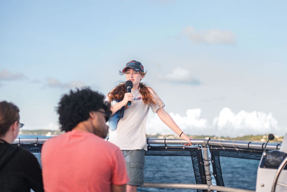 Woman speaking into microphone on boat deck with ocean background and blue sky.