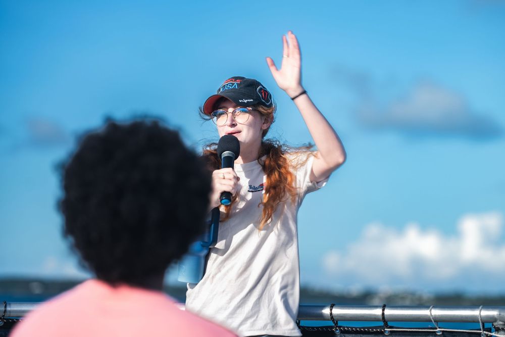 Young woman with red hair speaking into microphone, raising hand outdoors under blue sky.