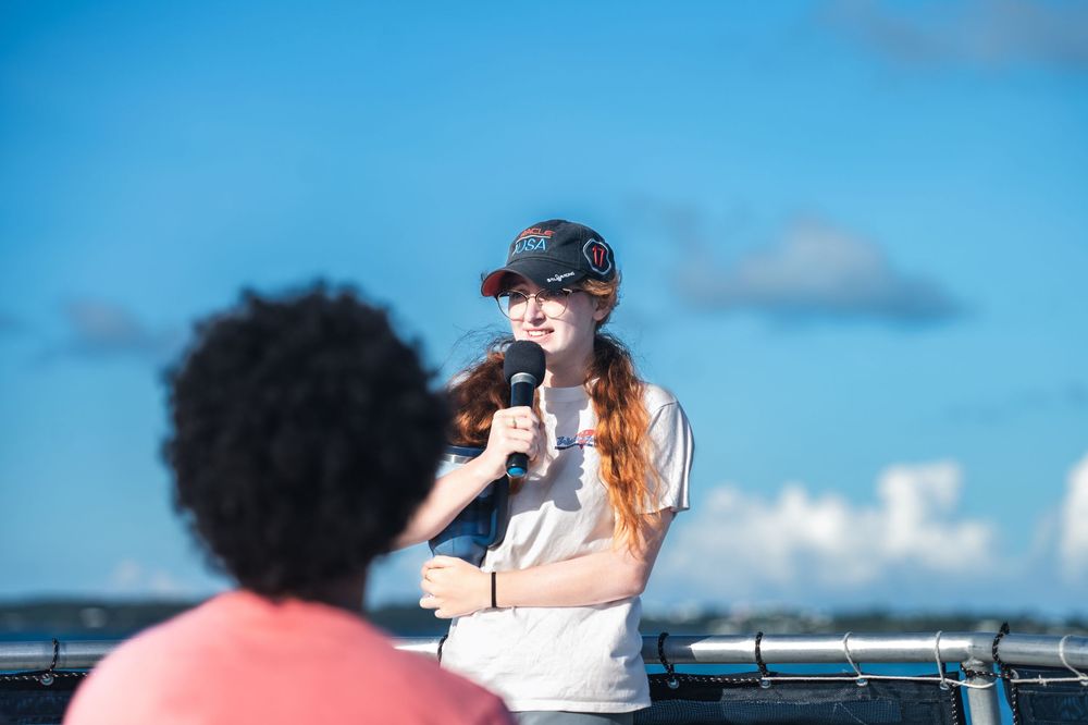 Young woman in cap speaks into microphone outdoors under blue sky at an event.