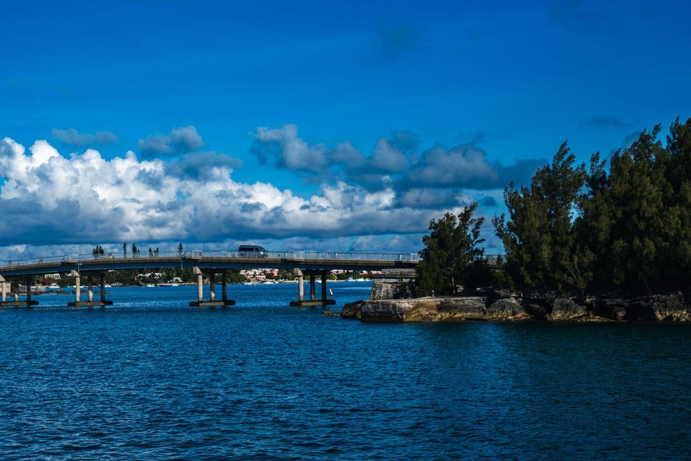A scenic bridge spans calm blue waters under dramatic cloudy skies, flanked by lush green trees on a sunny day.