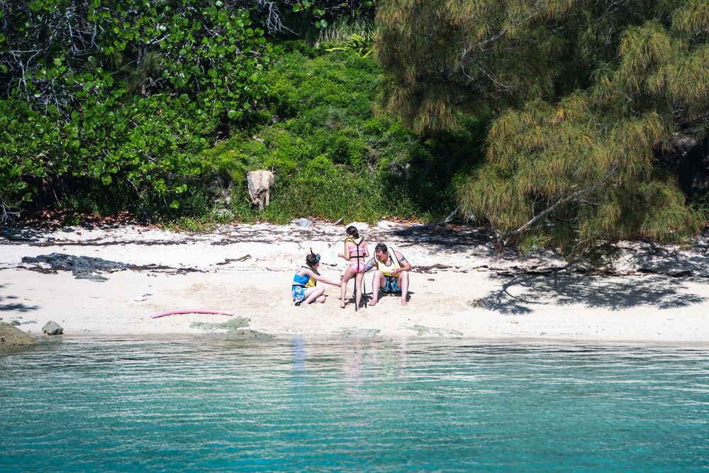People relaxing on a tropical white sand beach surrounded by turquoise water and lush green trees.