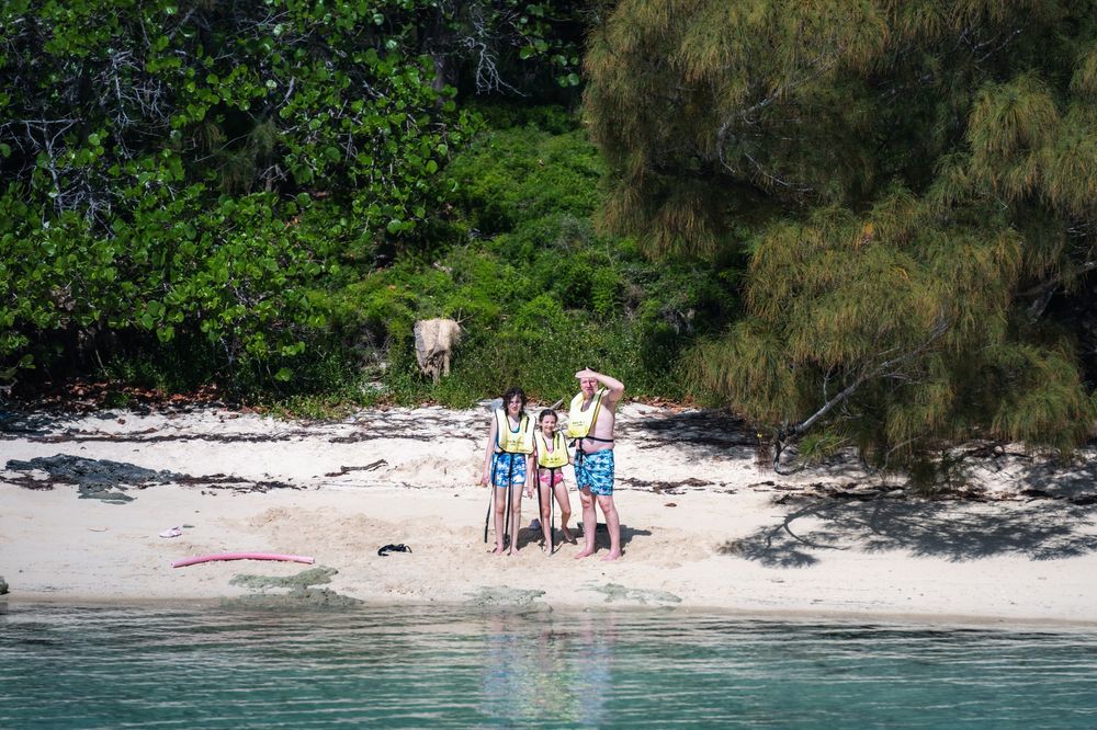 Family of three posing on a tropical white sand beach surrounded by lush green trees and clear turquoise water.
