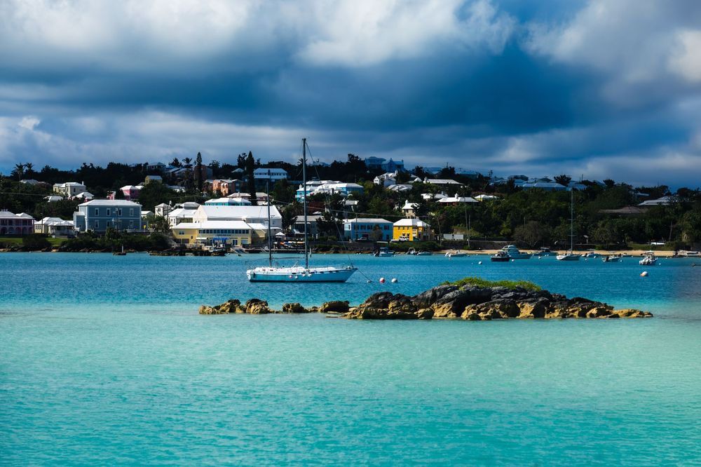Turquoise waters surround a rocky islet near a colorful coastal town under dramatic storm clouds in Bermuda.