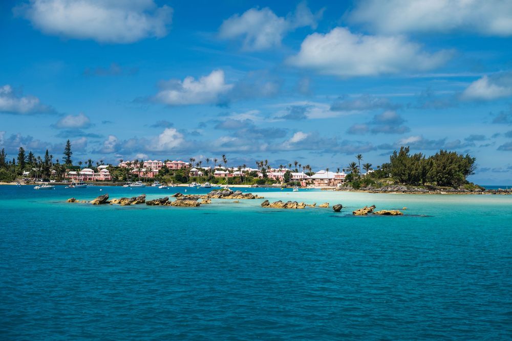Turquoise waters surround a rocky shoreline with colorful buildings and lush trees under a partly cloudy blue sky.