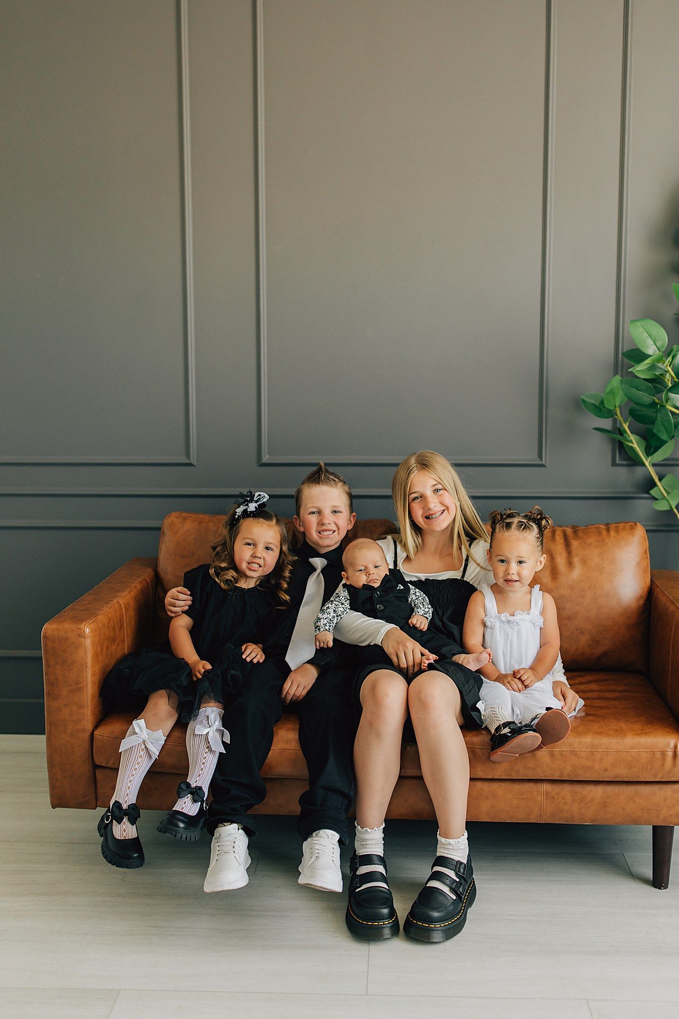 Blonde woman sitting on brown leather sofa with four young children in black and white outfits smiling.