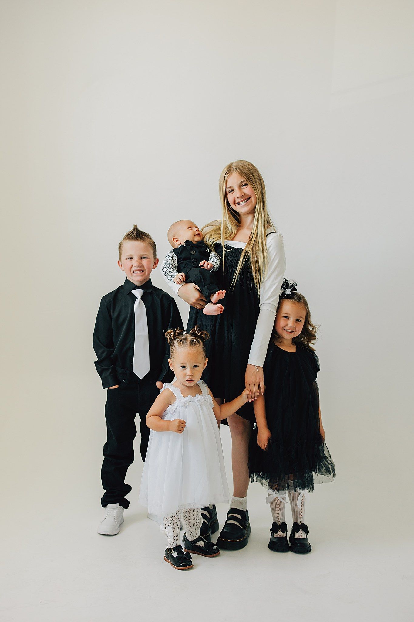 Smiling blonde woman holding a baby with three young children in black and white outfits posing together.