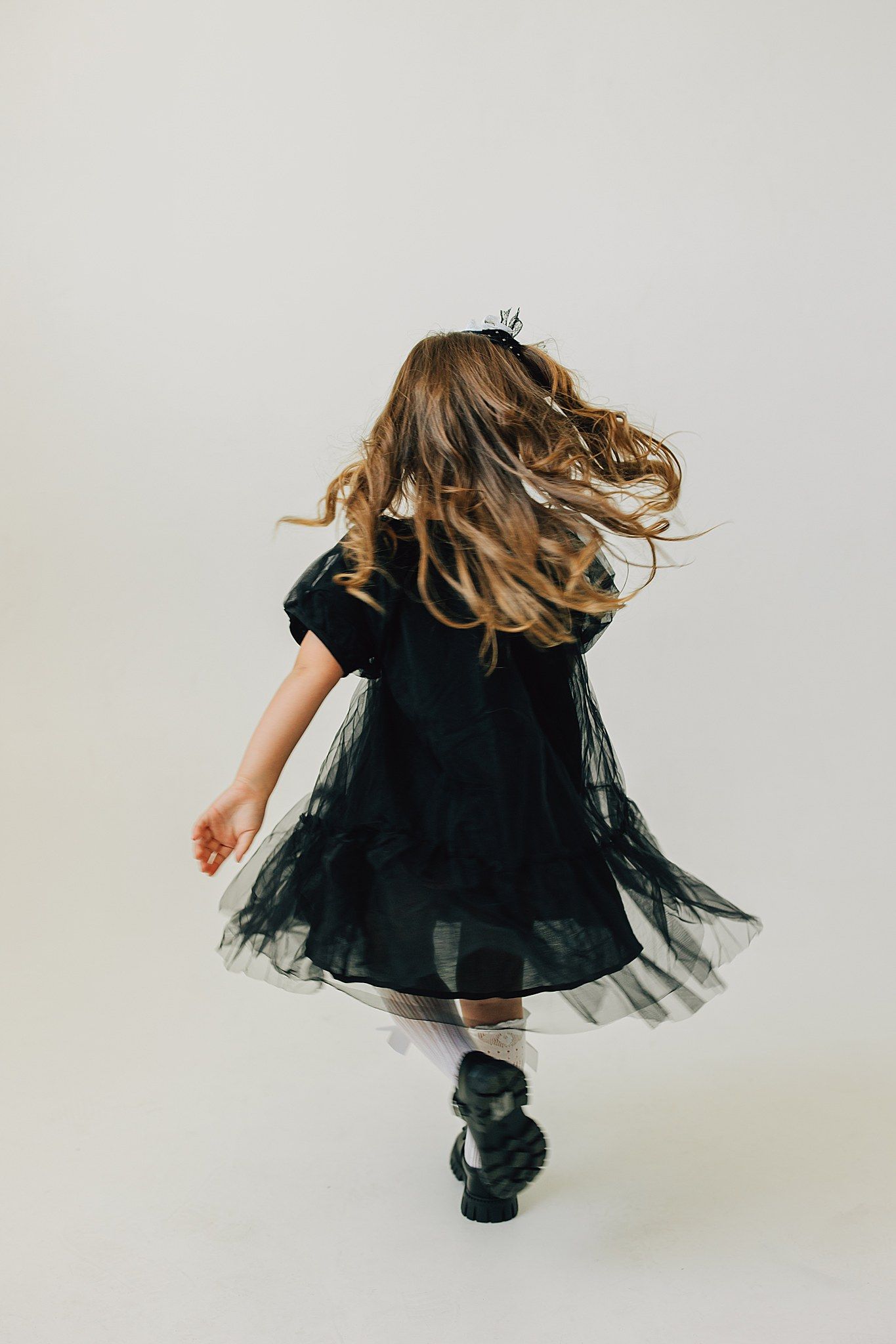 A young girl in a black tulle dress and crown twirls away from camera, her long hair flowing.