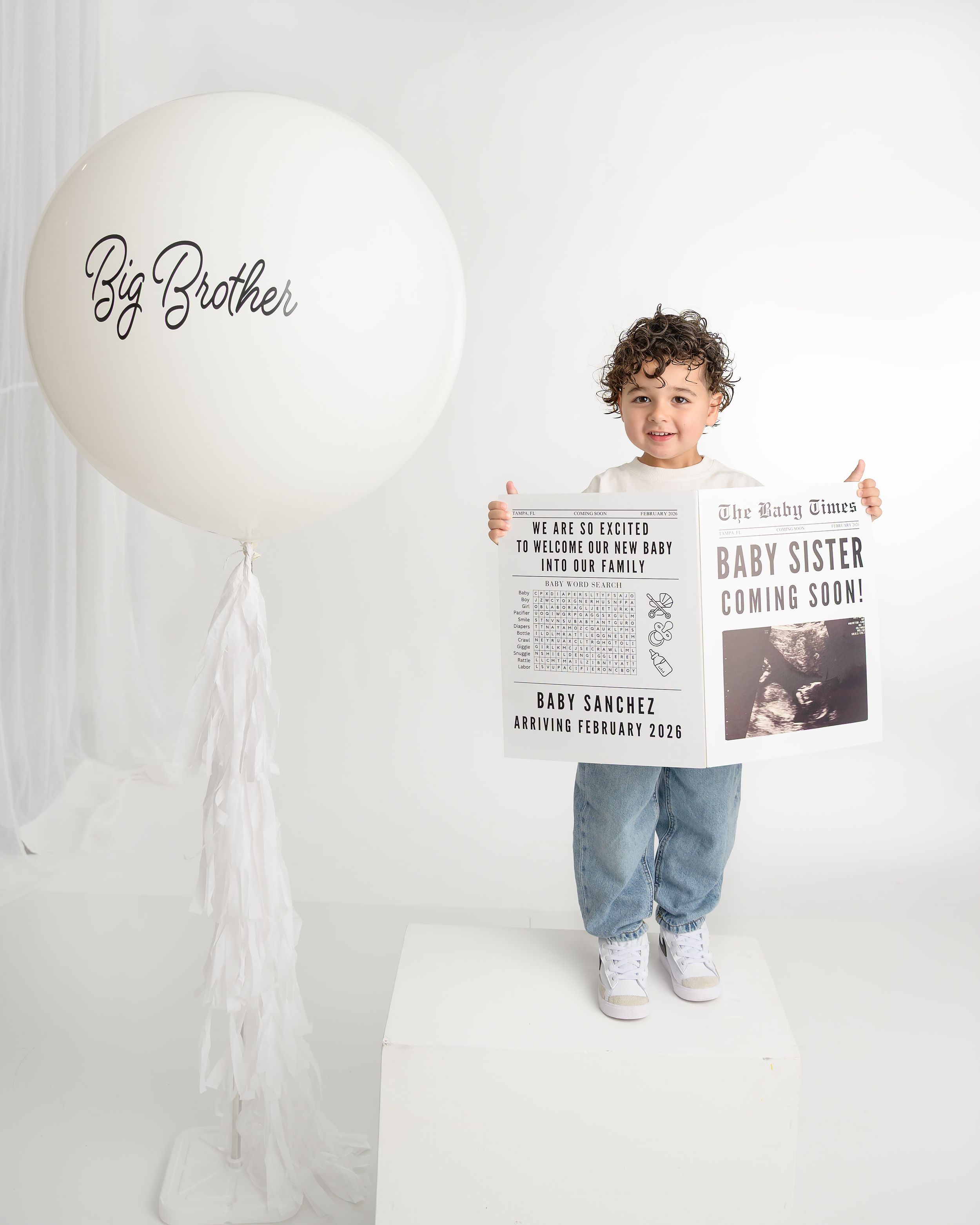 Toddler boy holding baby announcement newspaper beside a Big Brother balloon in white studio.