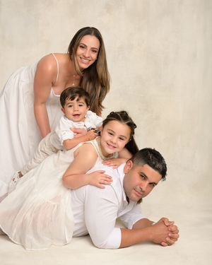 Family of four in white outfits posing together in a studio portrait, with parents and two young children smiling warmly.