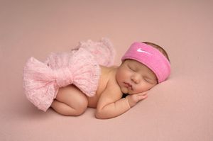 Newborn baby girl sleeping peacefully in pink tutu skirt and Nike headband on soft pink background.