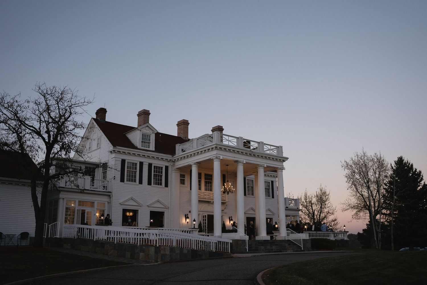 Grand white colonial mansion with tall columns and balcony glowing at dusk against a twilight sky.