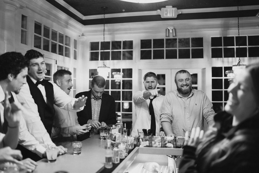 Black and white photo of groomsmen laughing and celebrating at a bar during a wedding reception.