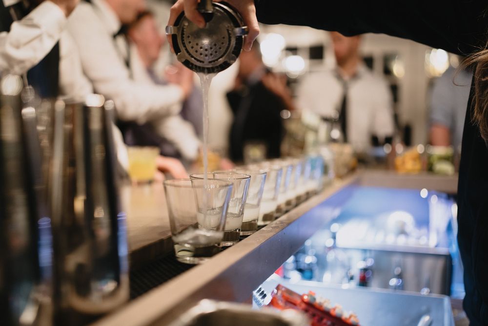 Bartender pouring cocktails into a row of glasses at a busy bar during an upscale event.
