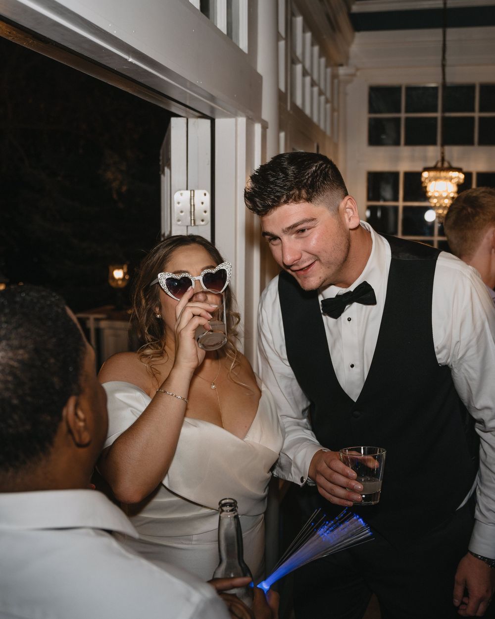 Bride in white dress with heart sunglasses laughing with groom in tuxedo vest at wedding reception.