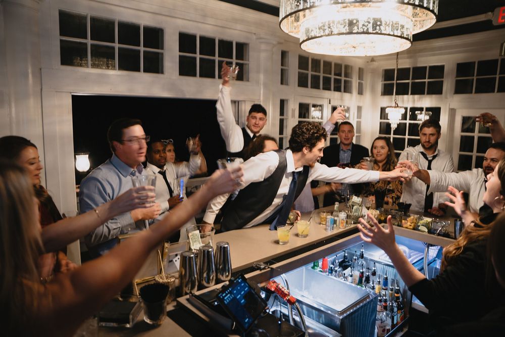Energetic bartender serving drinks to a lively crowd at an elegant indoor bar with chandelier lighting.