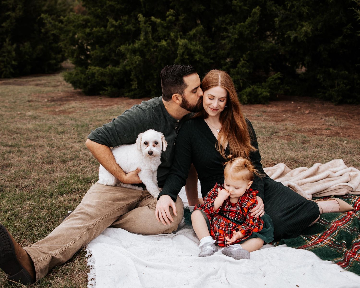 Family sitting on blanket outdoors with white dog, man kissing woman's head, toddler in plaid outfit.