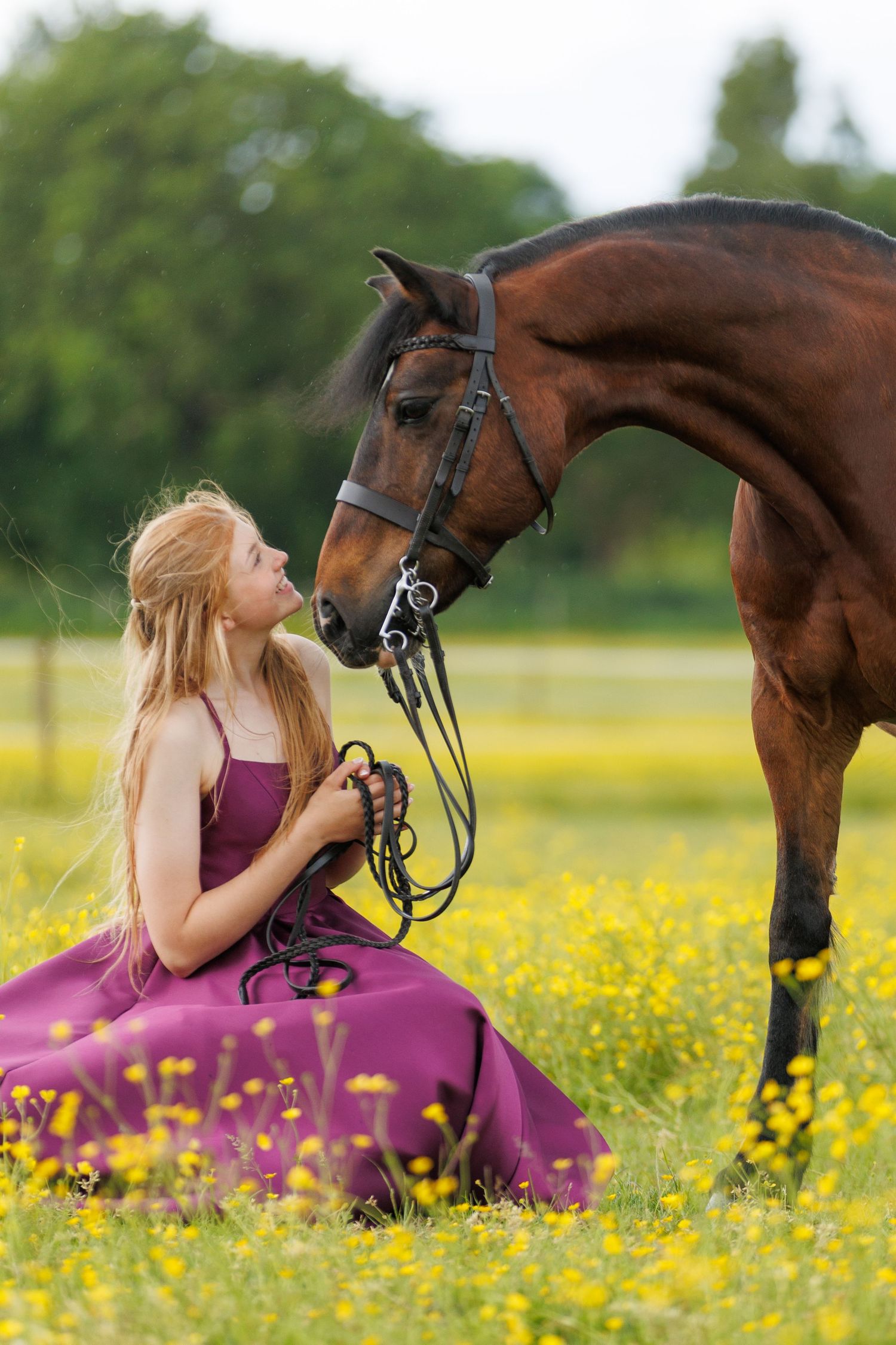 A smiling woman in a plaid shirt holds the reins of a brown horse beside a rustic stone wall outdoors.