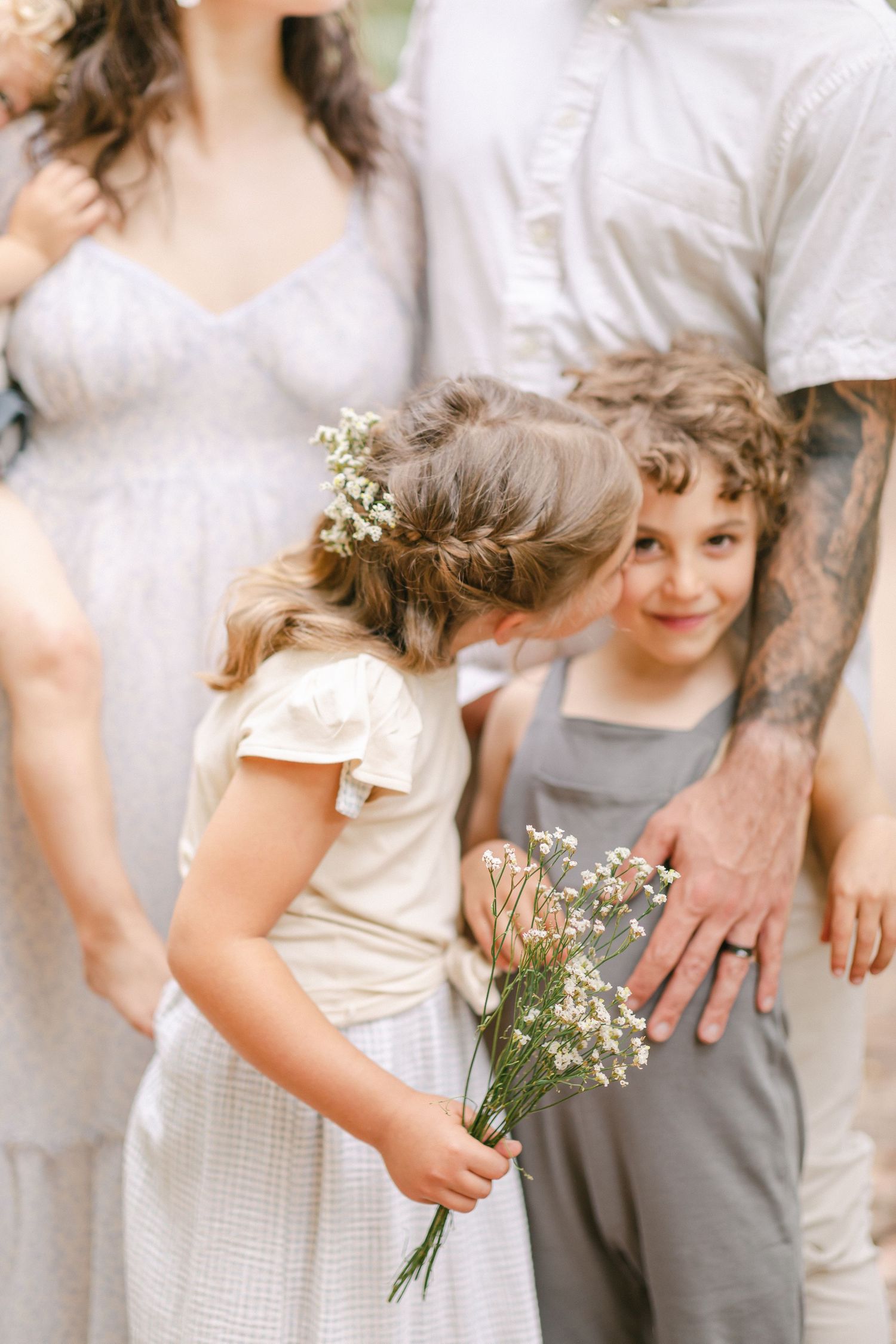 A flower girl holding wildflowers kisses a curly-haired boy at a rustic outdoor wedding ceremony.