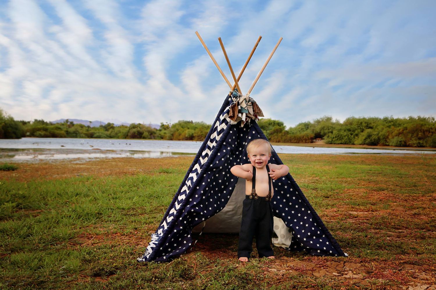Toddler boy in suspenders stands in front of a navy polka dot teepee on a grassy lakeside field under a blue sky with wispy clouds.