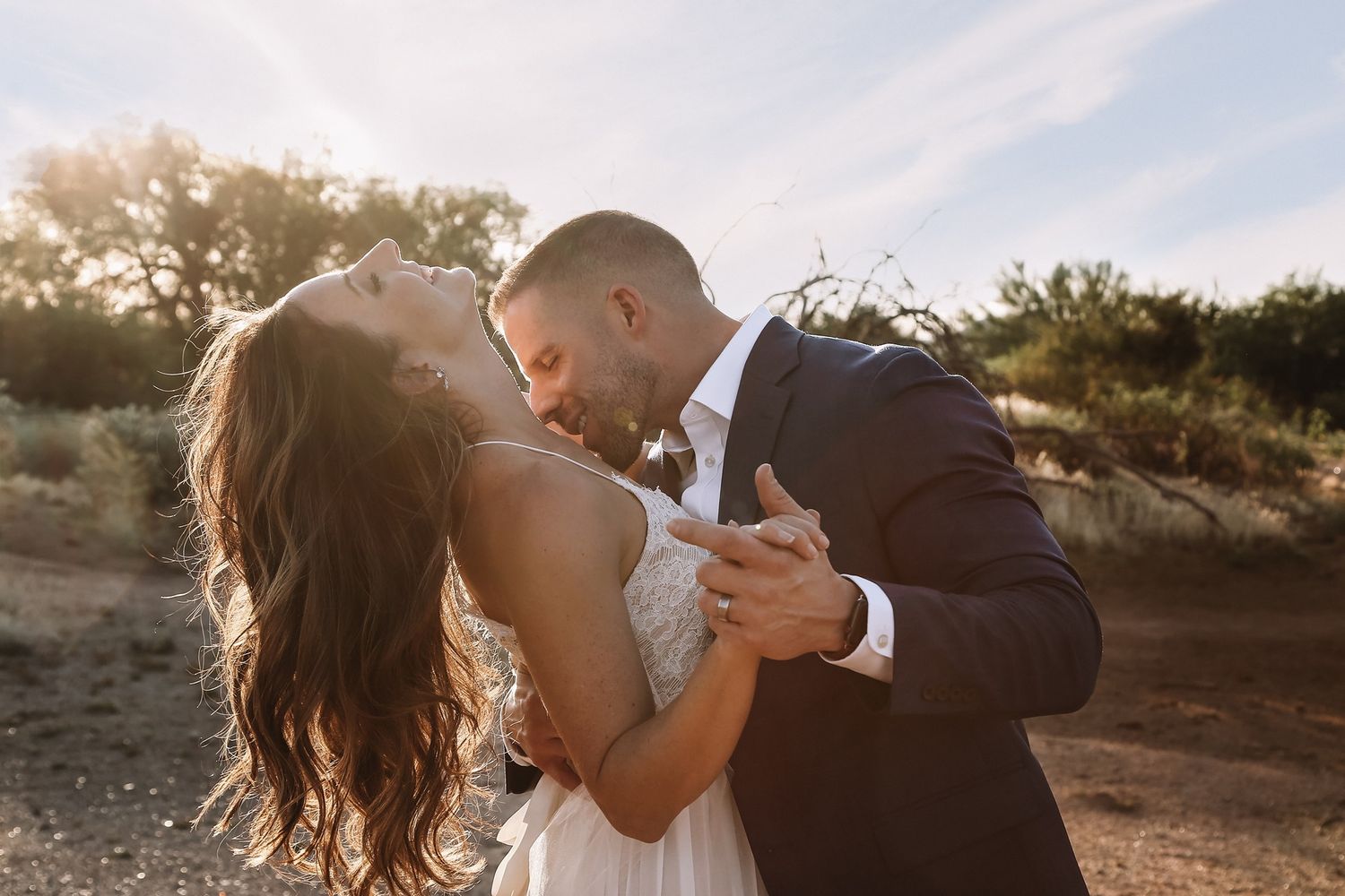 Bride and groom in a romantic dip kiss outdoors, with her long wavy hair flowing under warm golden-hour light.