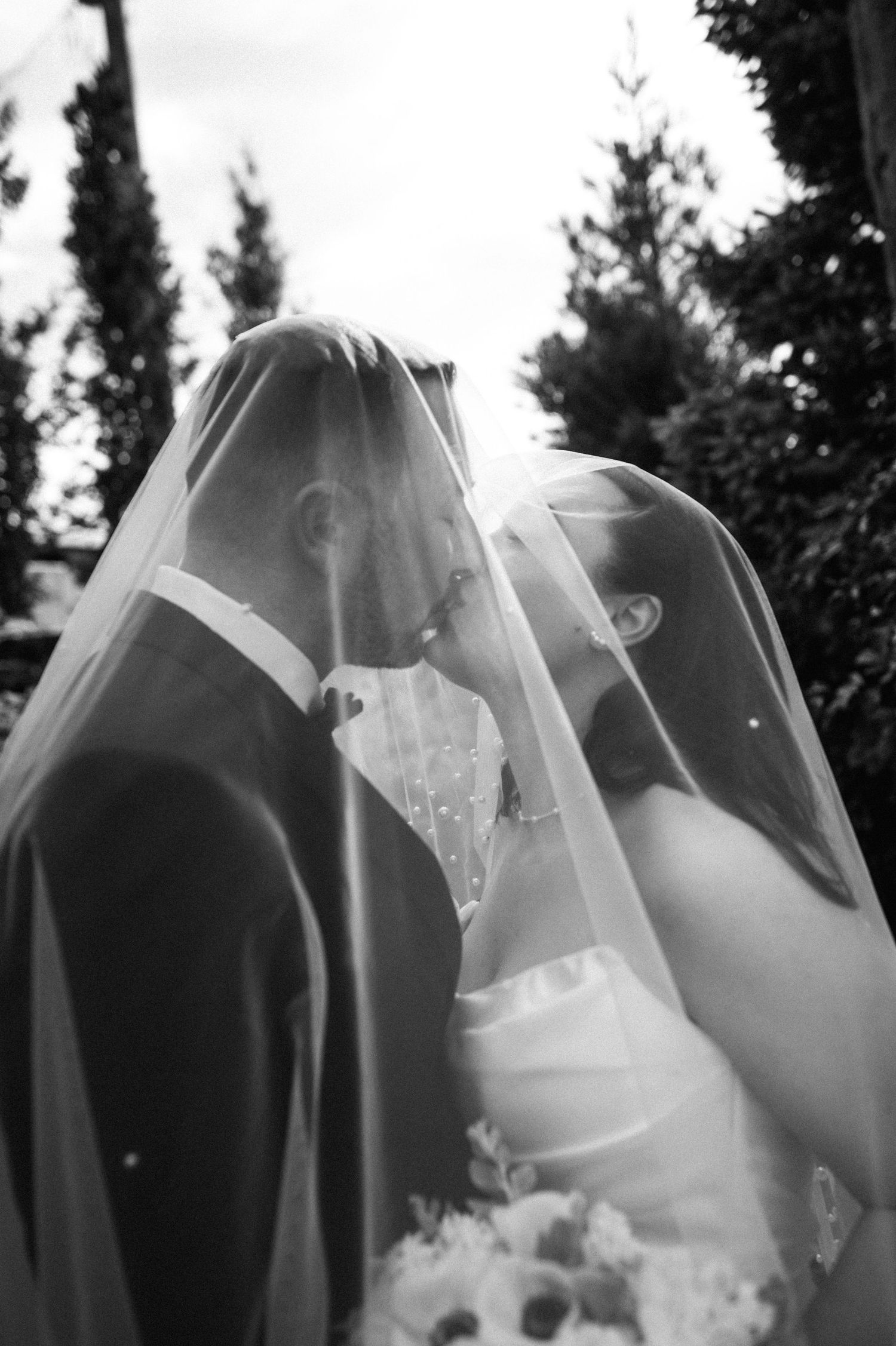 Black and white photo of bride and groom sharing a kiss under wedding veil outdoors.