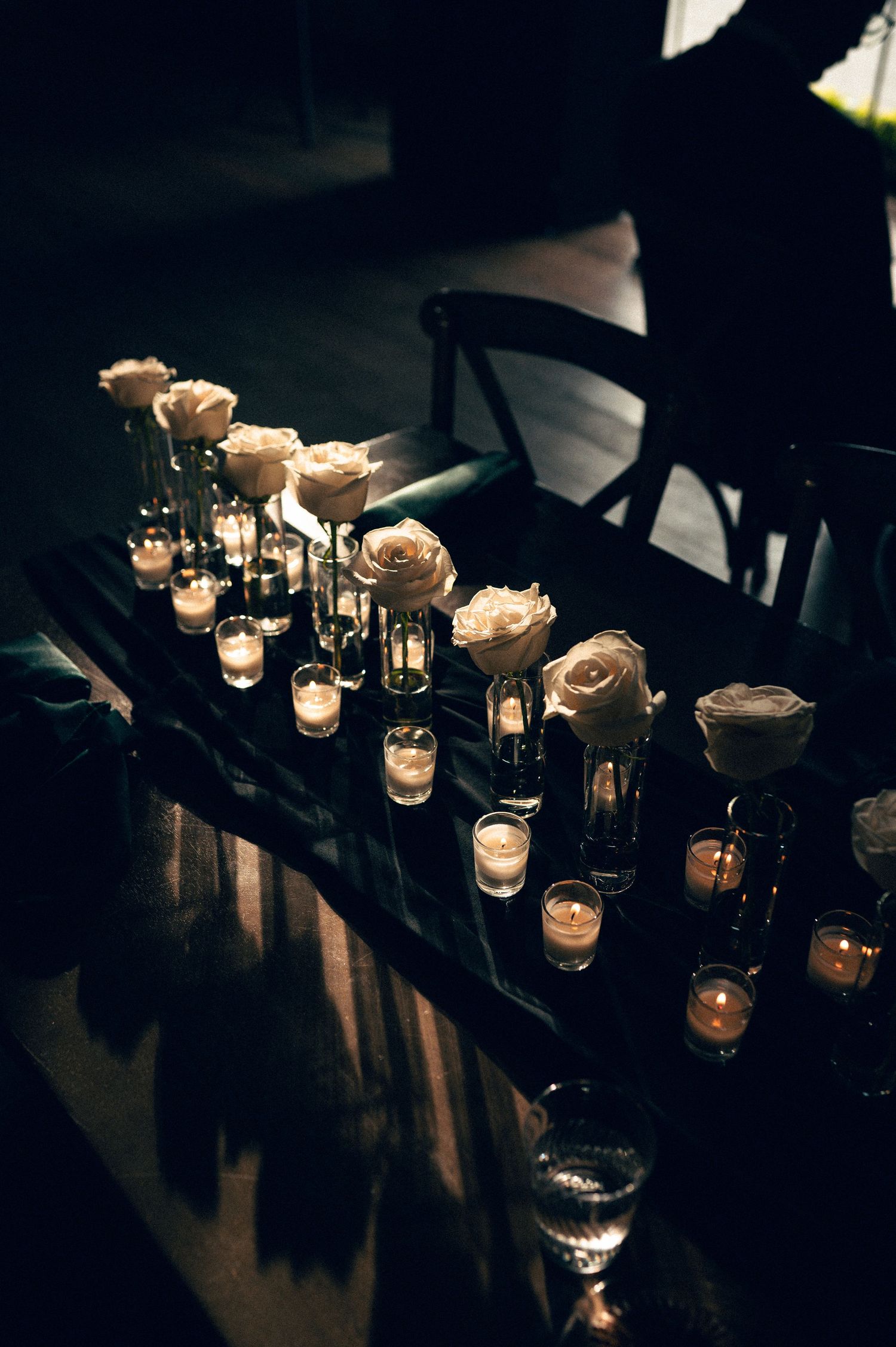 Elegant dark table with white roses in glass vases and glowing tea light candles creating romantic ambiance.