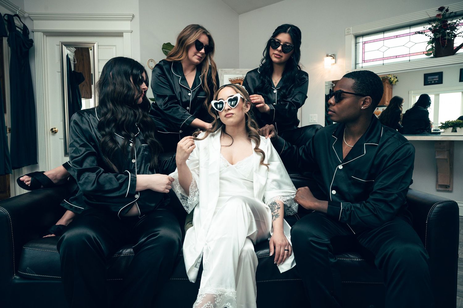 Bride in white robe surrounded by bridesmaids in black satin pajamas getting ready in a stylish room.