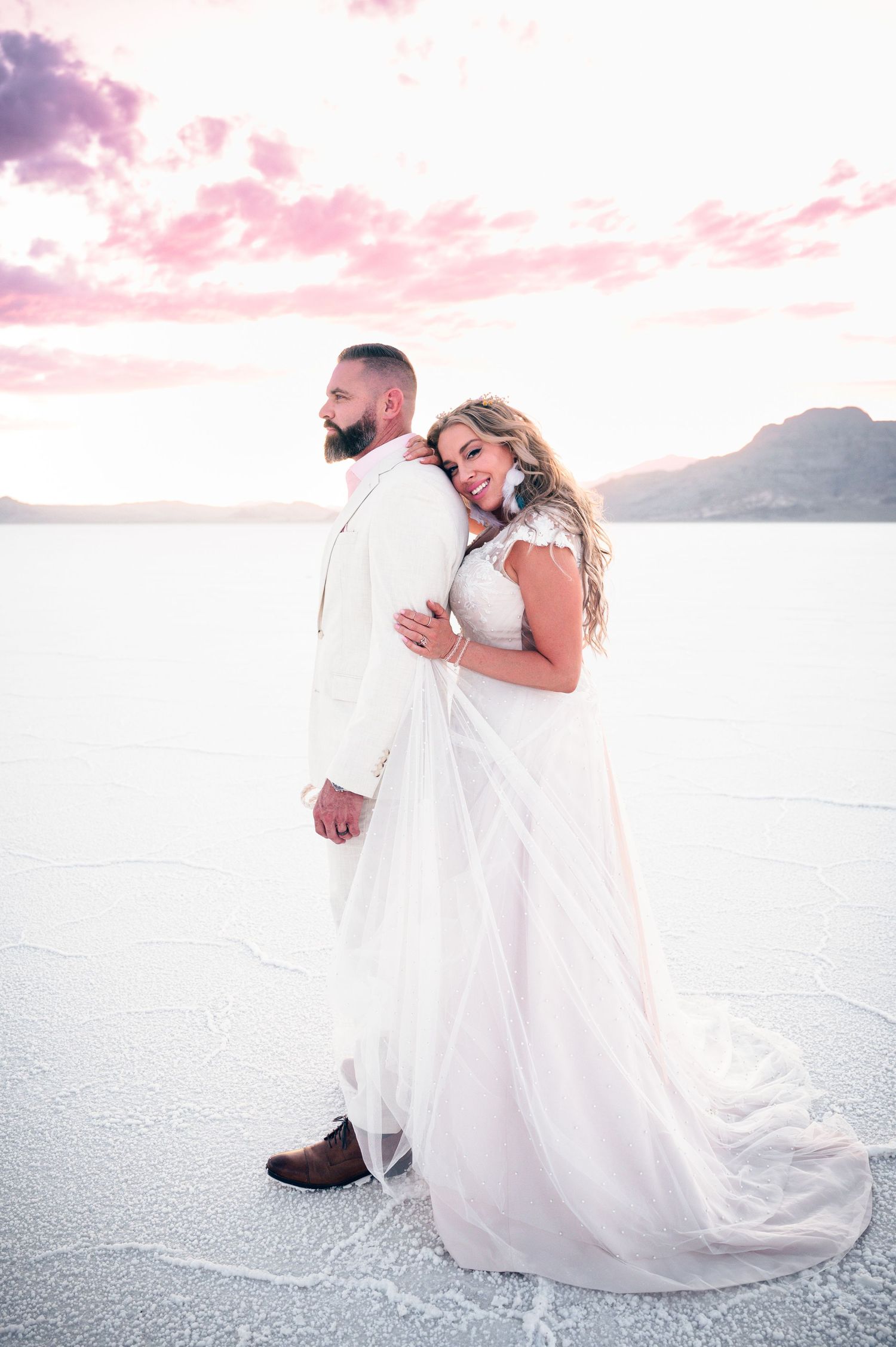 Bride in white gown embraces groom in white suit at Bonneville Salt Flats during a pink sunset wedding photo.