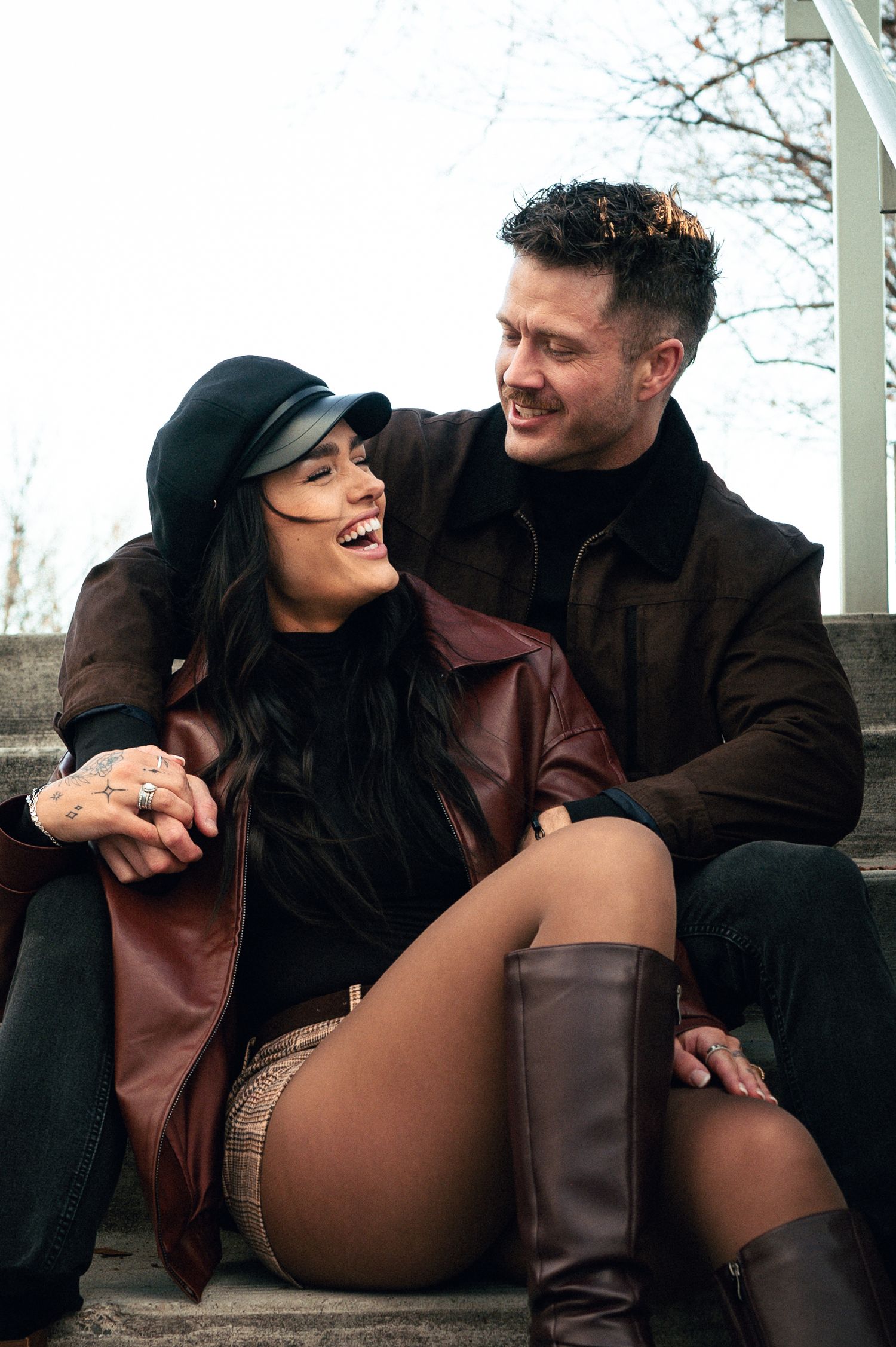 Happy couple sitting on steps outdoors, woman in brown leather jacket and black cap laughing with man in dark coat.