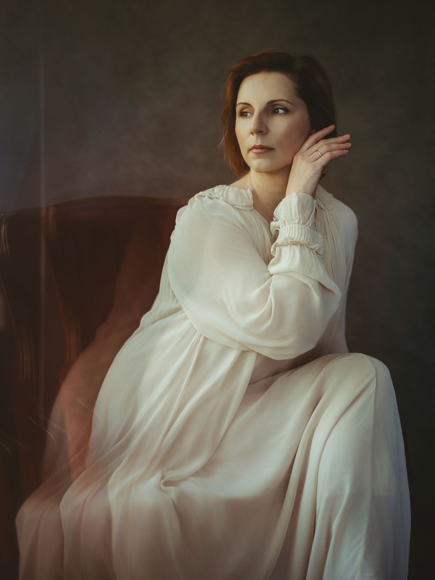 A woman in a black coat and top hat holds white wildflowers to her face with eyes closed against a dark background.