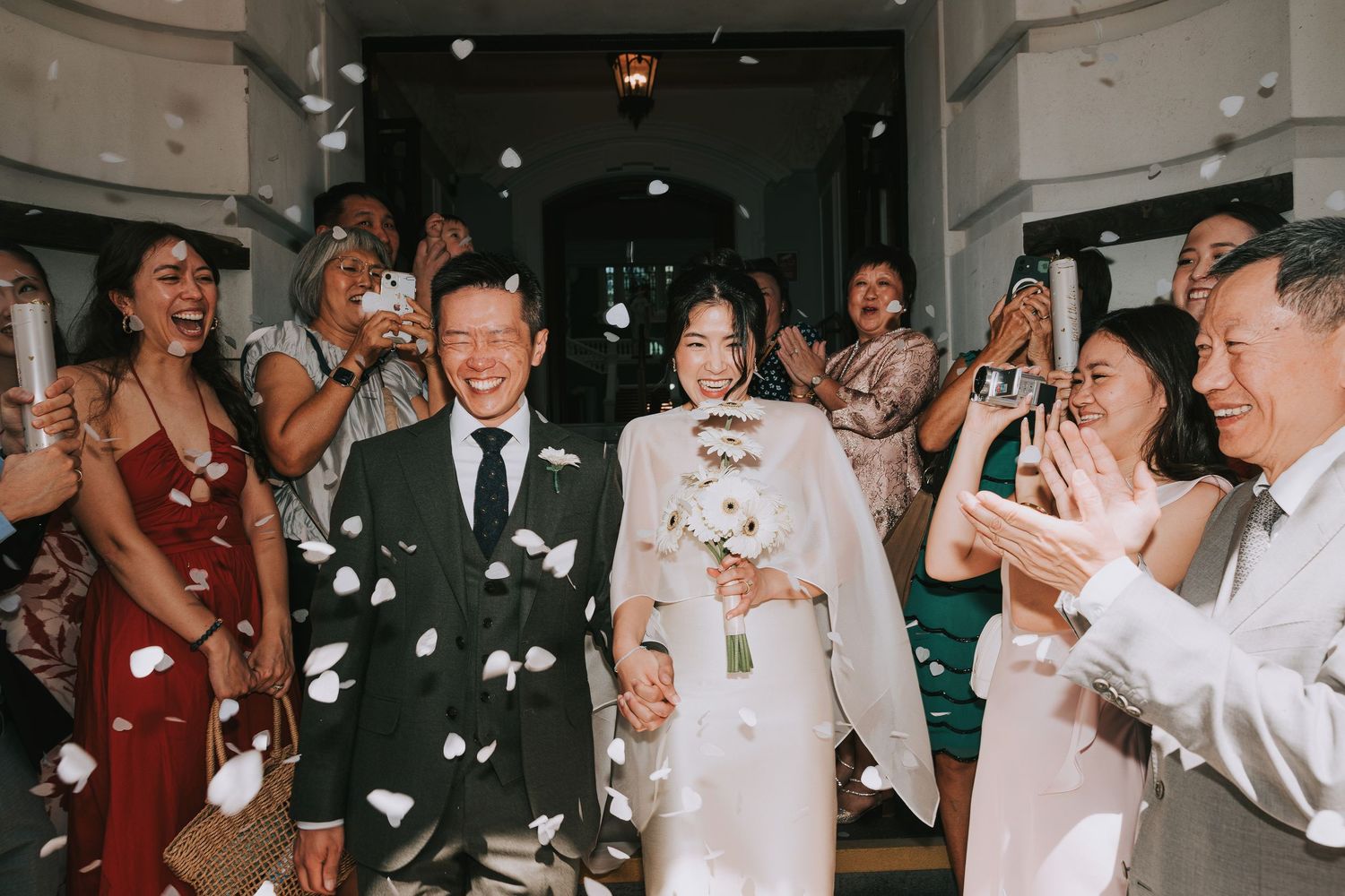 Newlywed couple walks through petal toss surrounded by cheering guests at elegant wedding ceremony exit at Woolwich Town Hall.