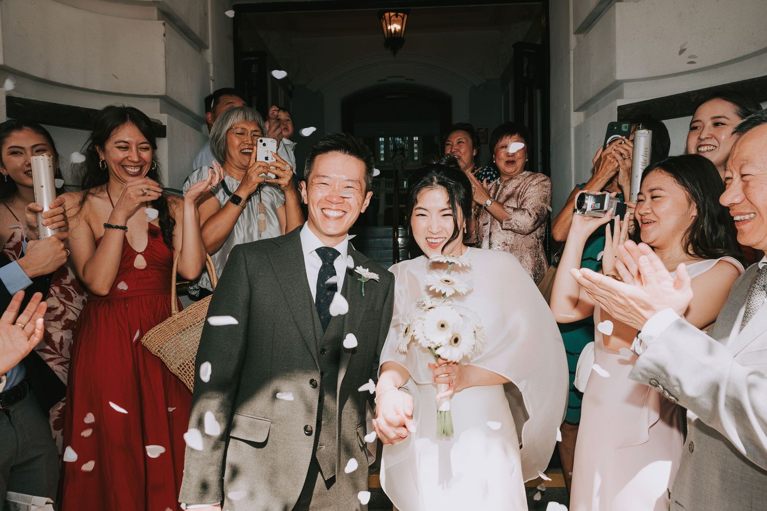 Joyful newlywed Asian couple walks through confetti shower as guests celebrate their wedding ceremony exit.