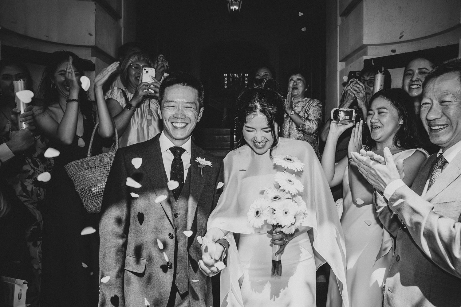 Black and white photo of a joyful couple walking through a flower petal toss at their wedding ceremony.