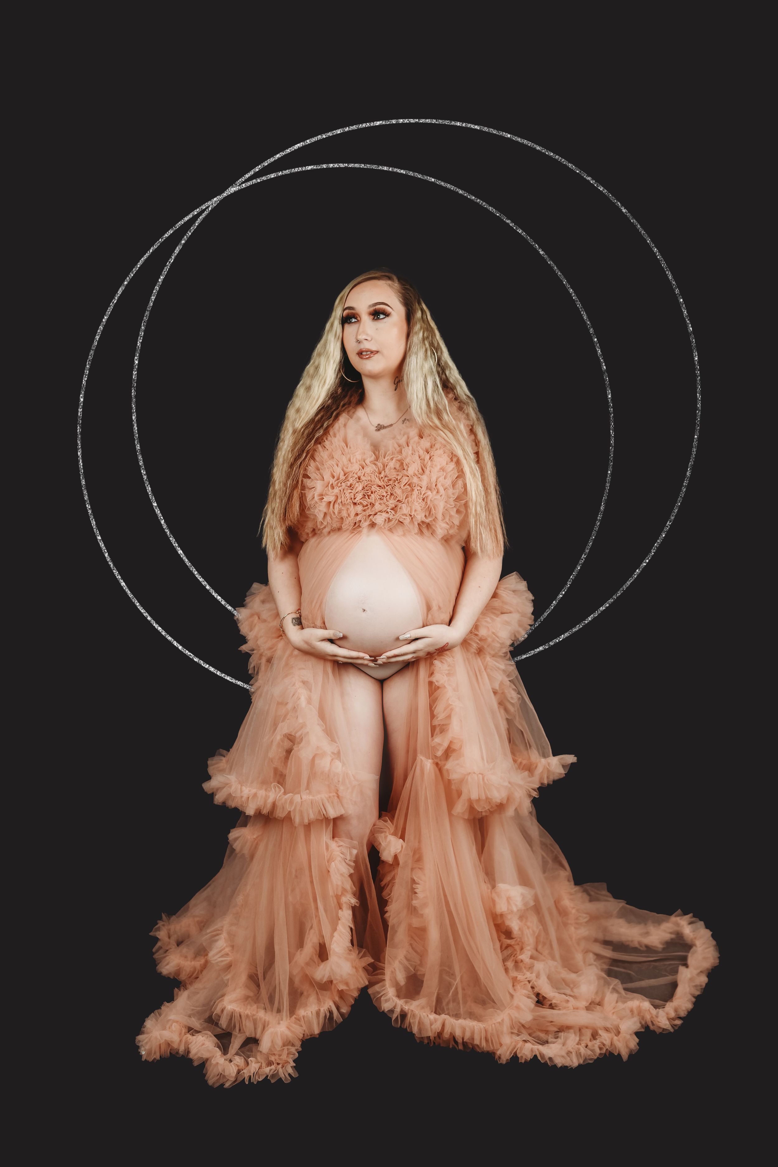 Pregnant woman in peach tulle robe cradling baby bump against black background with circular light design.
