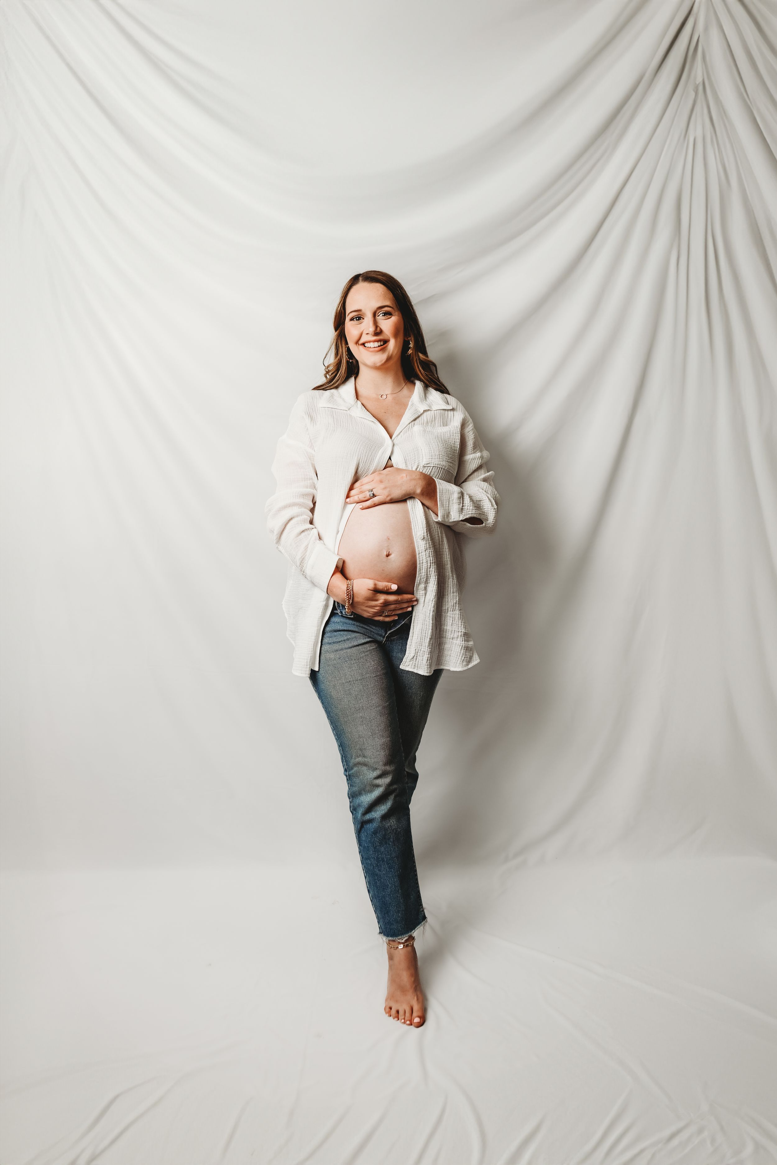 Smiling pregnant woman in open white shirt and jeans cradling bare baby bump against white draped backdrop.