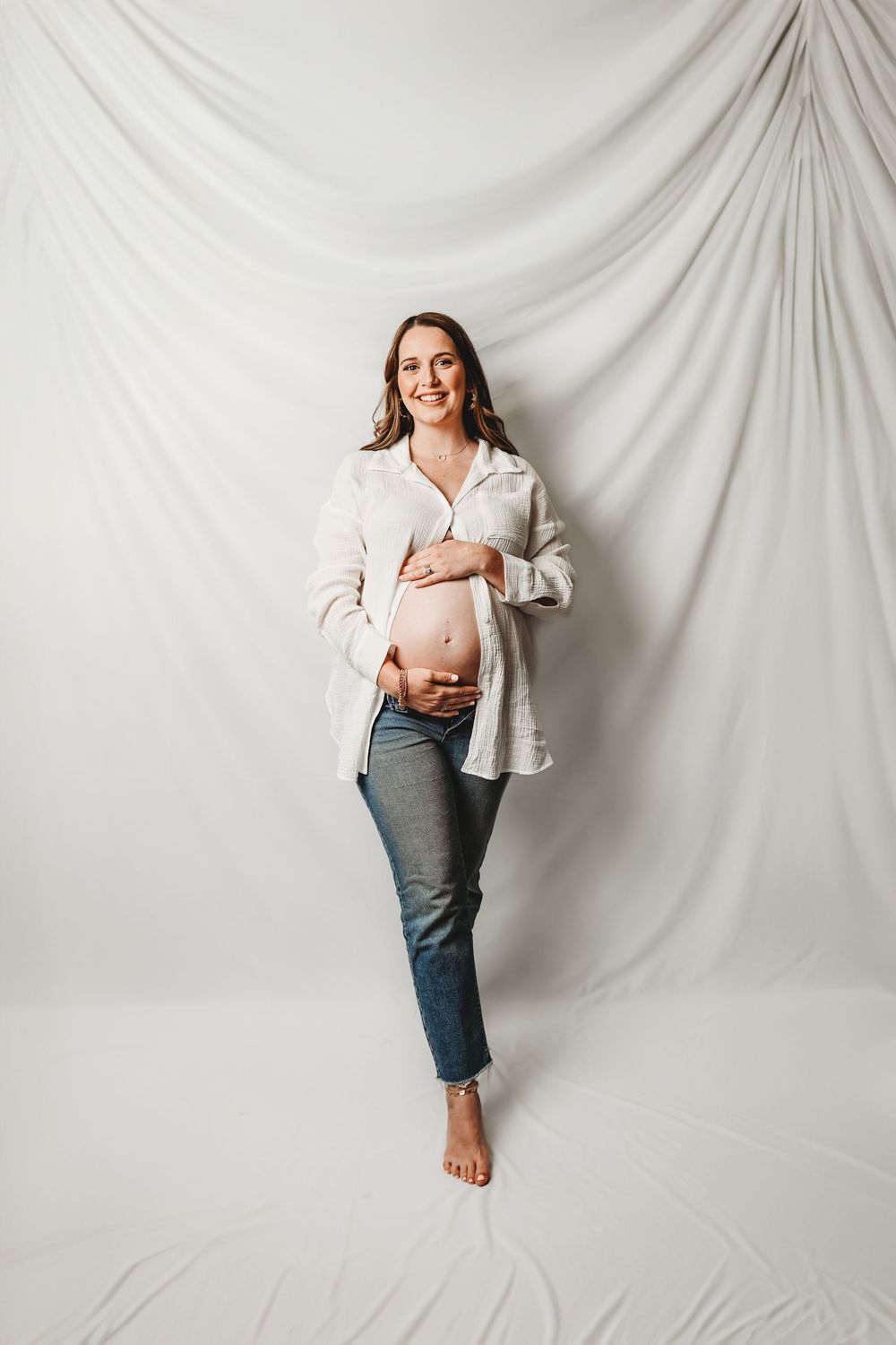 Smiling pregnant woman in open white shirt and jeans cradling bare baby bump against white draped backdrop.