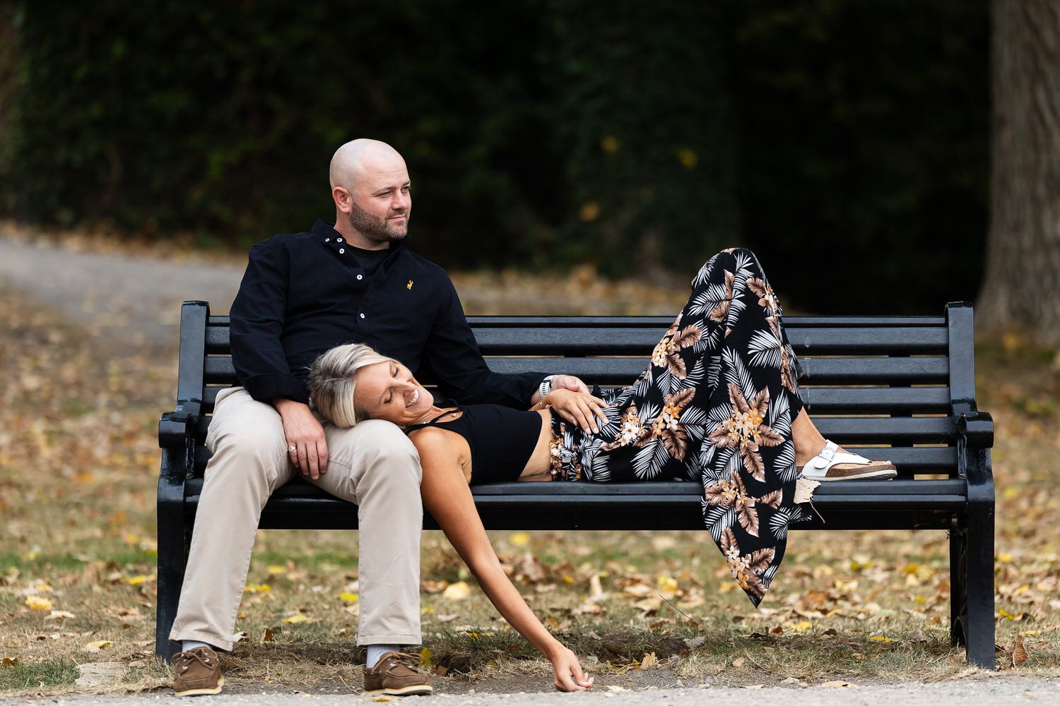 Couple relaxing on a park bench, man sitting upright while woman reclines with legs up, surrounded by autumn foliage.