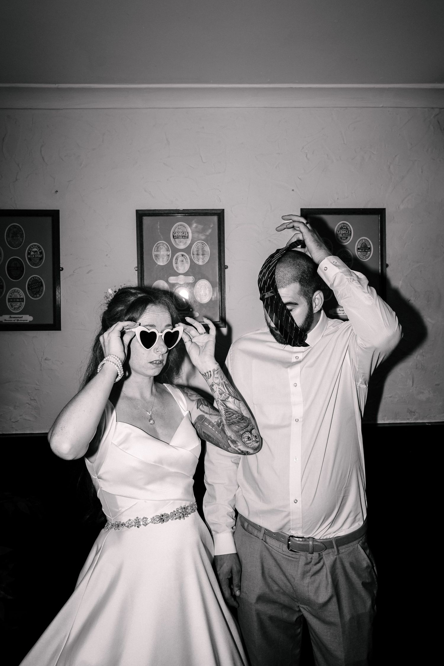 Black and white photo of a bride in heart-shaped sunglasses and tattooed groom dancing joyfully at their wedding reception. Couple Shot's