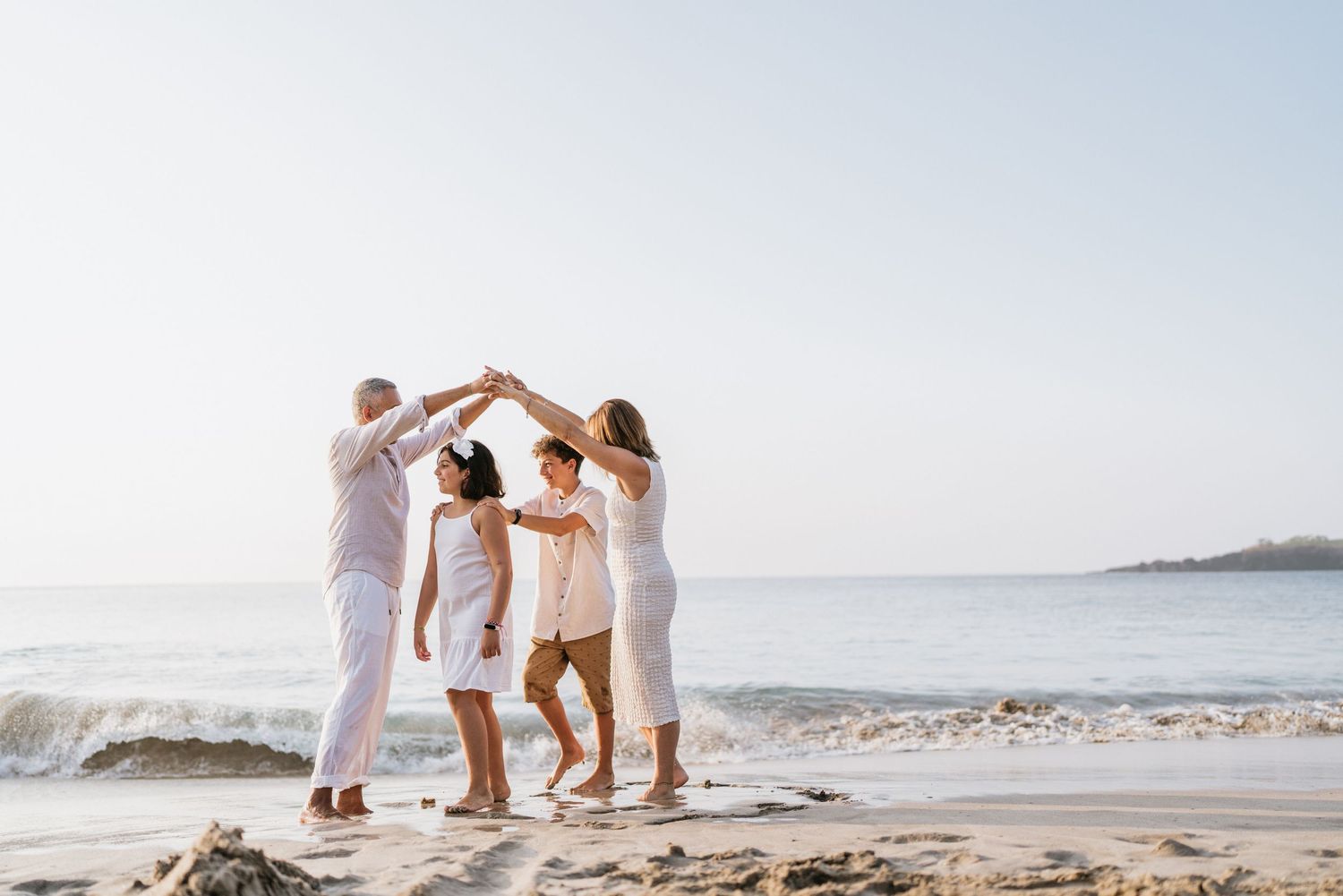 Family group playing and dancing together on sandy beach with ocean waves in background.