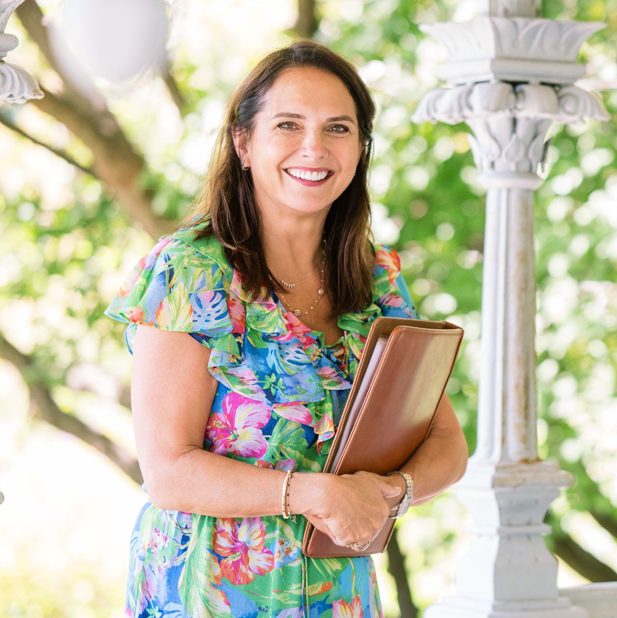 Smiling woman in floral dress holding a brown folder, standing outdoors near a white column.