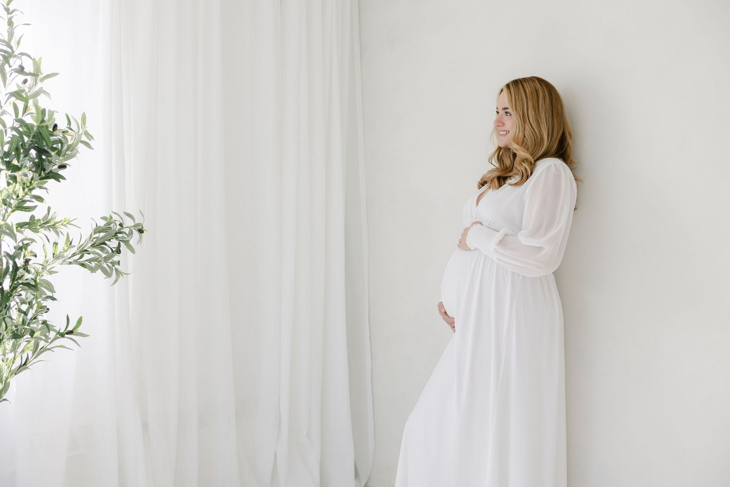 Pregnant woman in white dress cradling baby bump, standing by white curtains in bright minimalist room.