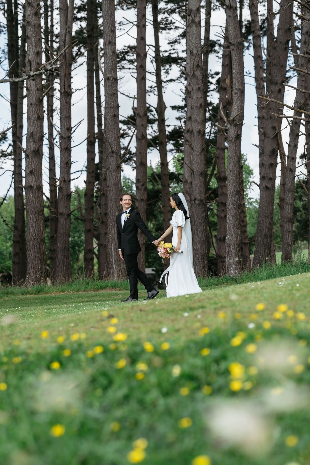 Wedding couple walking hand in hand on green grass with tall pine trees in the background.
