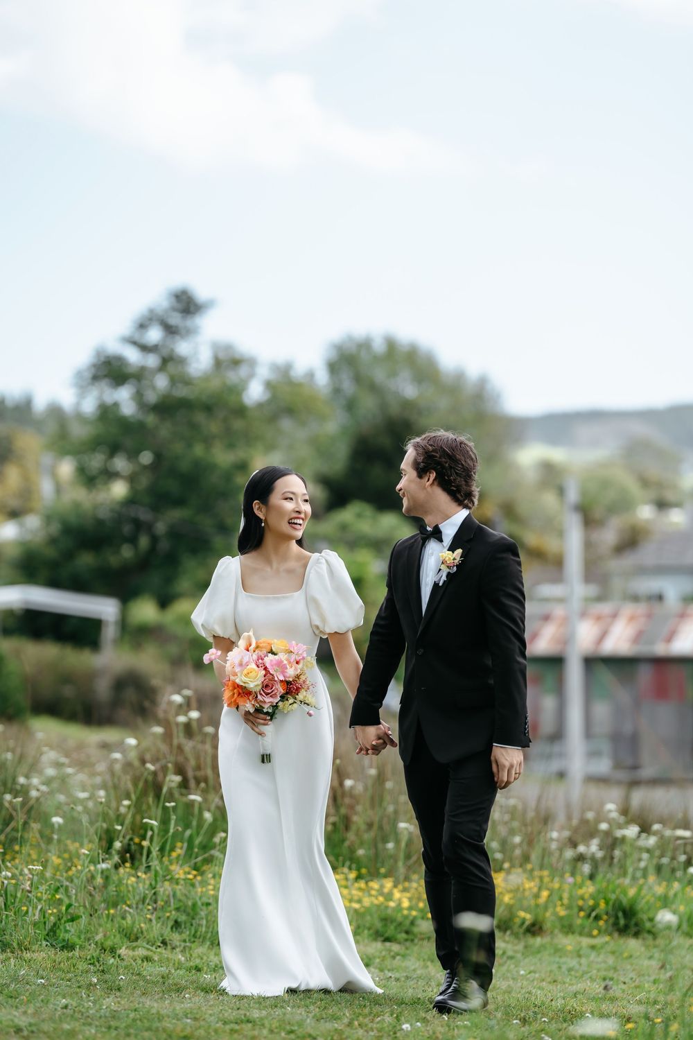 Bride and groom holding hands walking through a wildflower meadow, bride in white gown holding colorful bouquet.