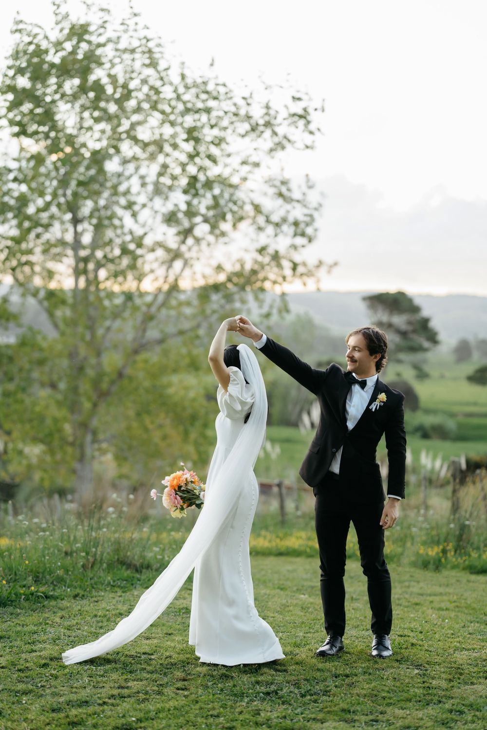 Bride and groom dancing outdoors on lush green grass at sunset, bride in white gown with long train, groom in black tuxedo.