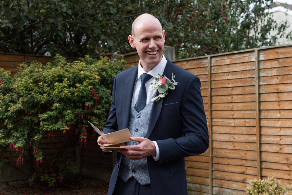 Smiling bald groom in navy suit with floral boutonniere holds a card outdoors near wooden fence.