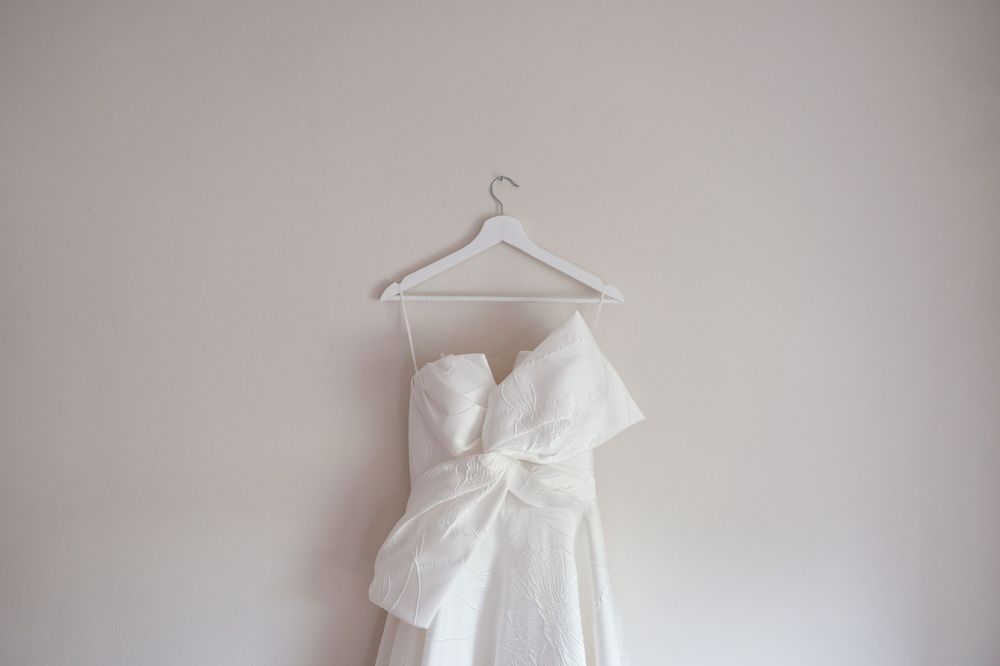 White strapless wedding dress with large bow detail hanging on a hanger against a neutral wall.
