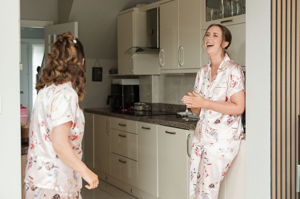 Two women in floral pajamas laughing together in a modern kitchen during a bridal morning preparation.