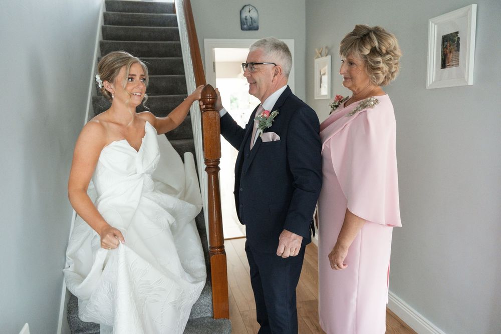 Bride in white gown holding hands with older man in navy suit beside woman in pink dress near staircase.