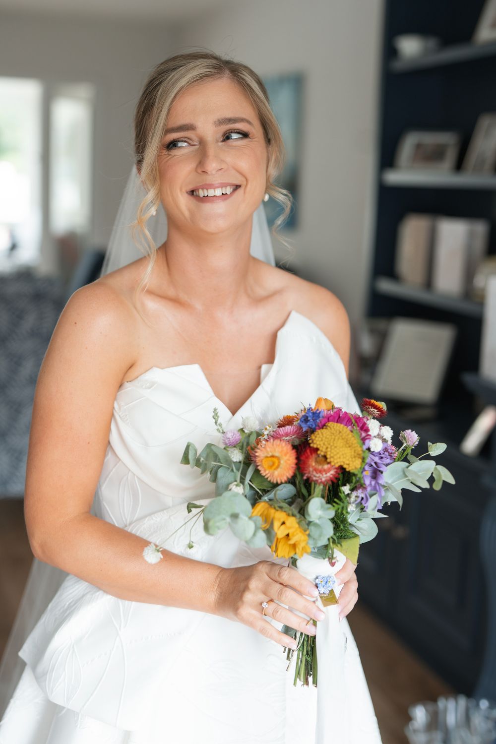 Smiling bride in strapless white gown holding colorful bouquet of orange, yellow, and purple flowers indoors.
