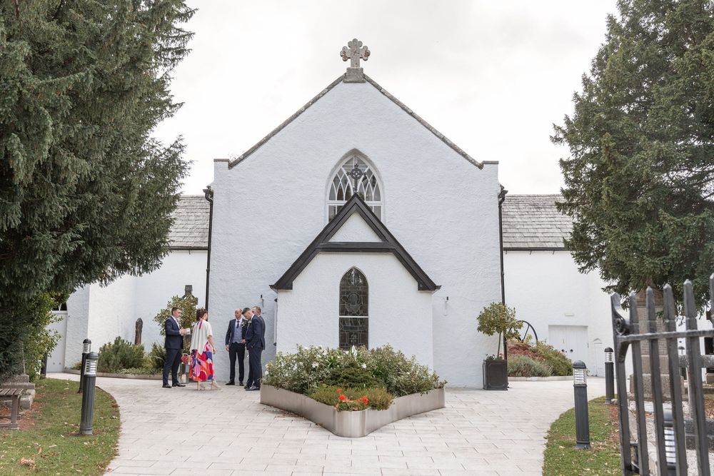 Wedding guests gather outside a charming white Gothic church with arched windows and a cross, surrounded by tall evergreen trees.